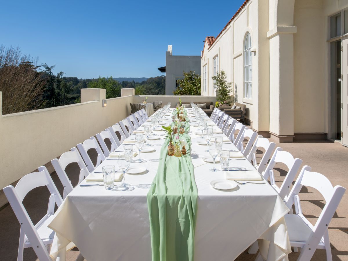 An outdoor terrace dining table set for a party, with white chairs, glassware, and a green centerpiece against a blue sky.