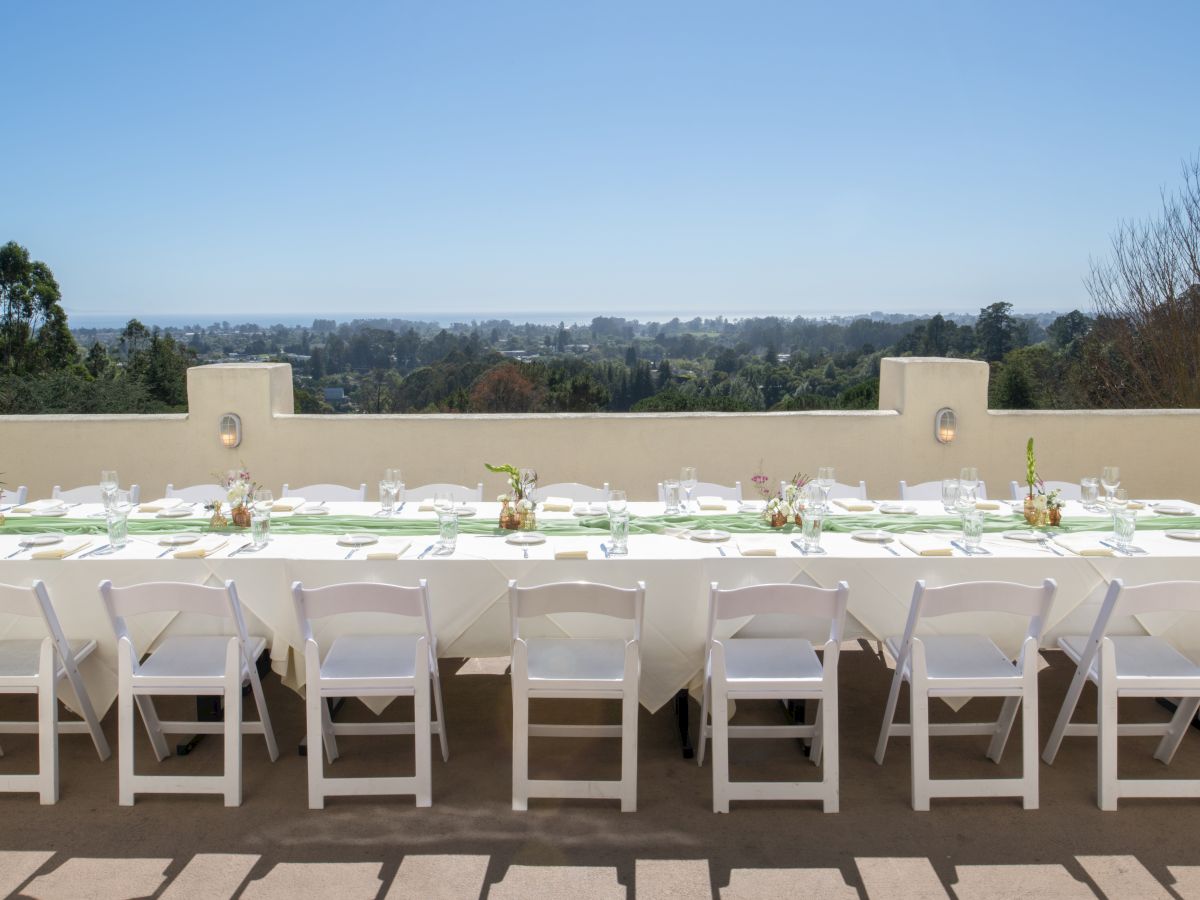 An outdoor terrace set for dining&mdash;white chairs and a table with glassware&mdash;overlooking lush hills under a clear blue sky.