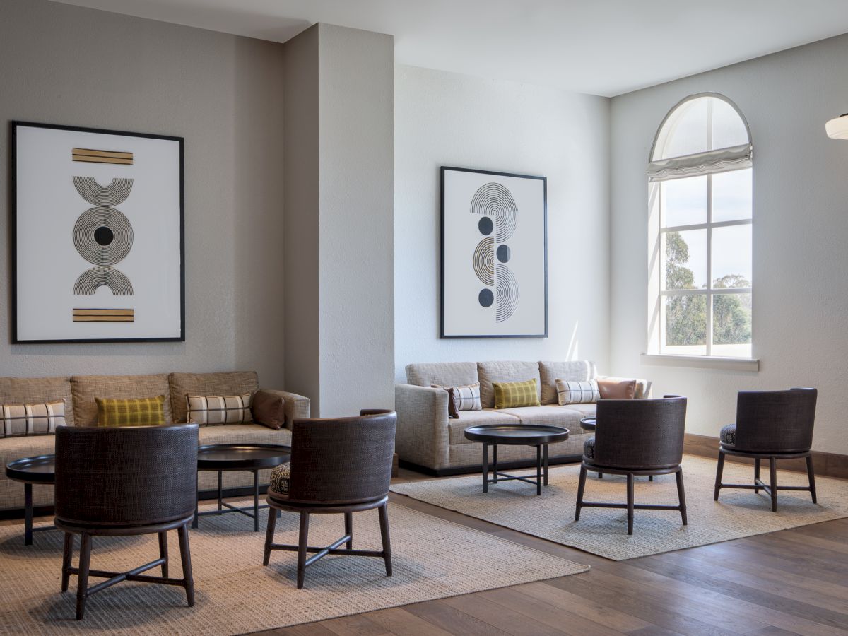 Bright modern living room with sofas, armchairs, coffee tables, framed abstract art, big window, and wall sconce lamp.