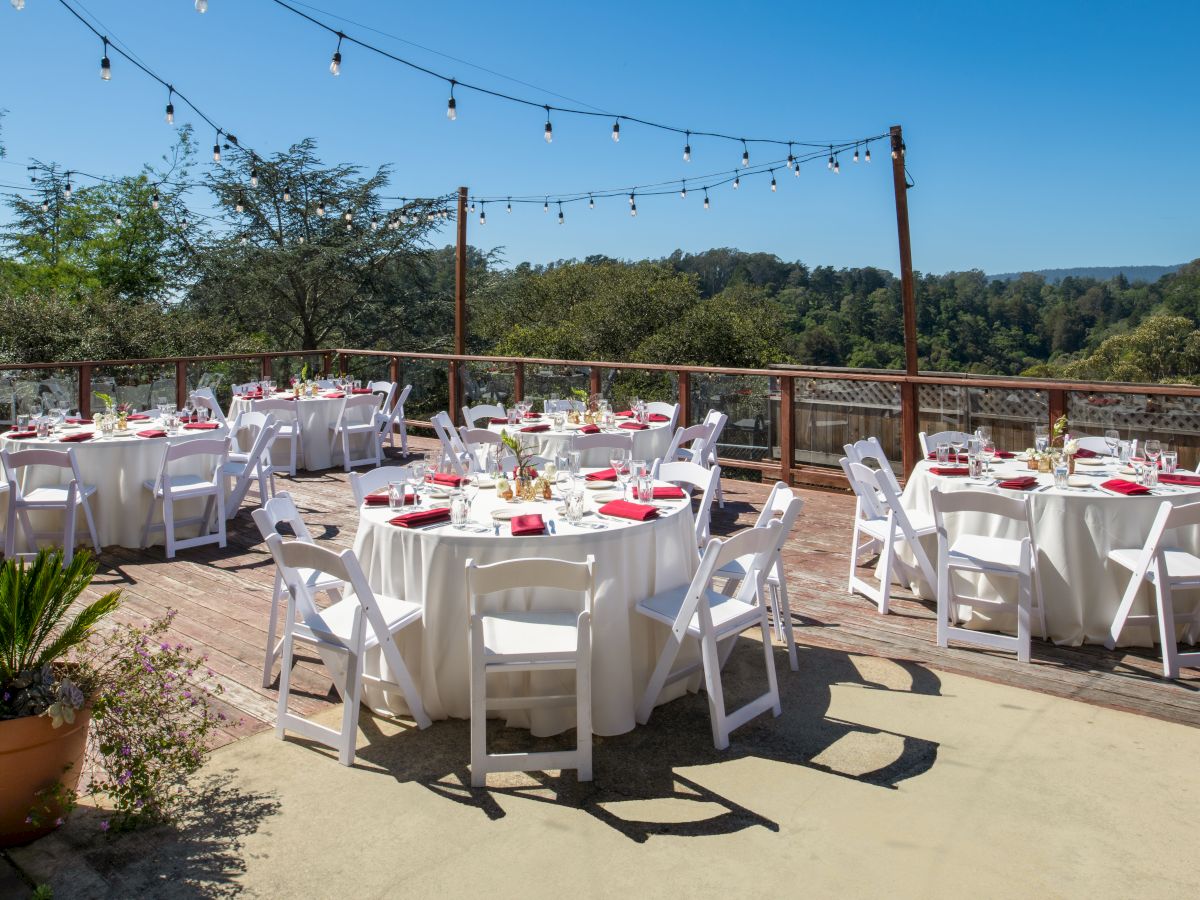An outdoor dining setup on a sunny deck: white chairs, covered tables with glassware, red napkins, and hanging string lights above a wooden railing.