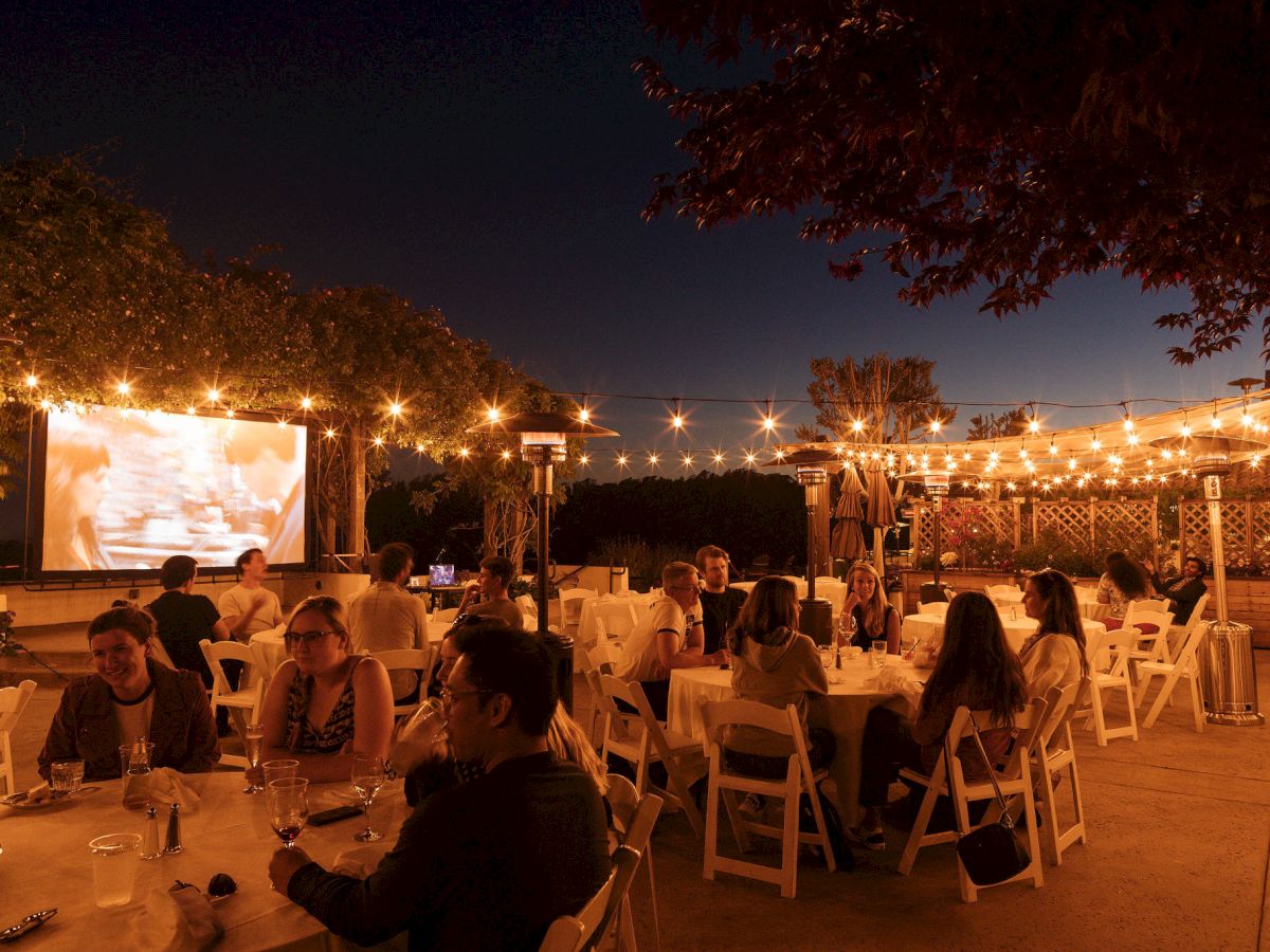 An outdoor dinner party with guests seated at tables under string lights, while a big screen plays on a warm evening.