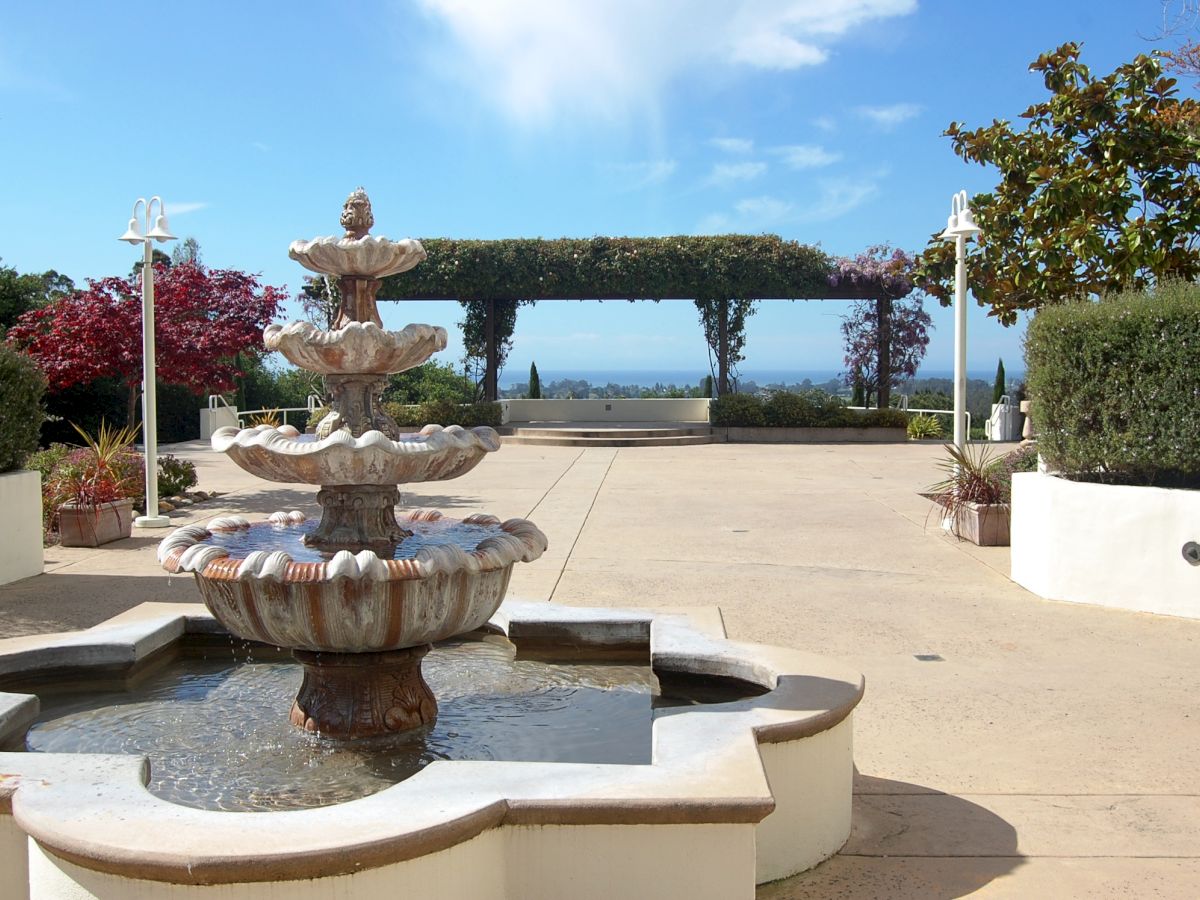 A landscaped courtyard fountain with a carved statue, surrounded by hedges and flowers, with trees and a distant ocean view.