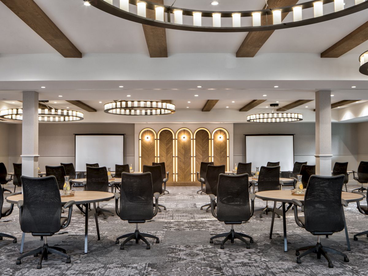A modern meeting room with round tables, swivel chairs, wood arches, and hanging ring chandeliers.