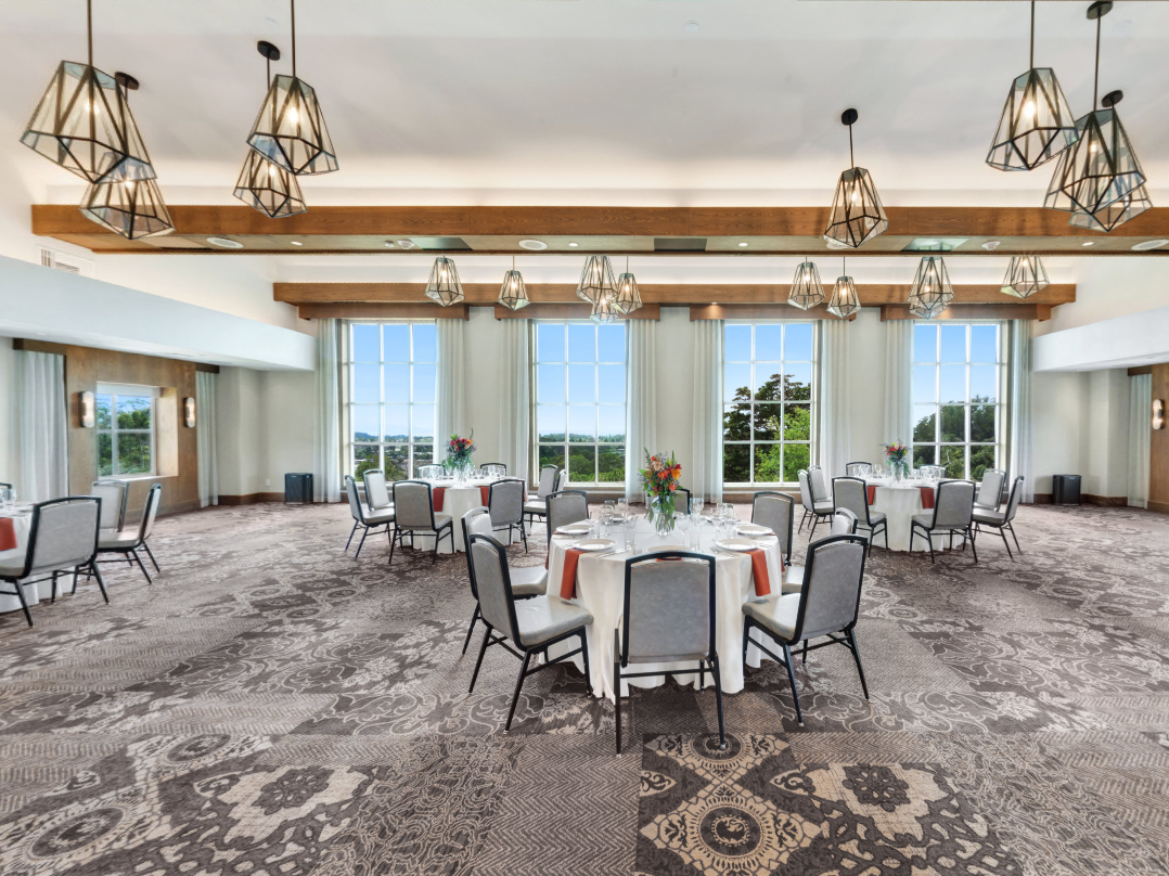 Bright event hall with round tables, grey chairs, floral centerpieces, and geometric pendant lights under large windows.