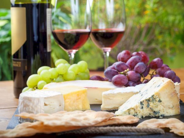 The image shows a wine and cheese platter with different cheeses, green and red grapes, and crackers, accompanied by two glasses of red wine.