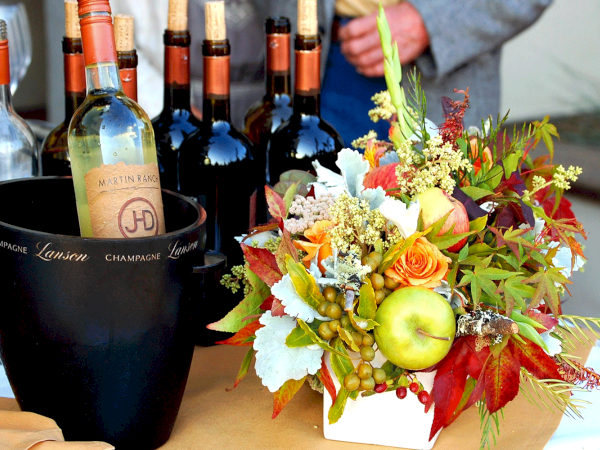 The image shows multiple wine bottles, a black ice bucket, and a colorful floral arrangement with fruits and flowers on a table.