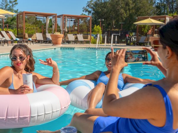 Friends relax by a bright outdoor pool, floating in inflatable rings and laughing while someone films on a phone.