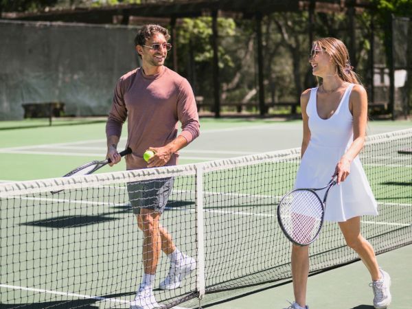 Two tennis players smile on a sunny court, one holding a racket and a green tennis ball while the other holds her racket.