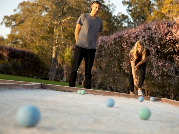 Two people outdoor playing bocce ball on a dirt court. The background has greenery.