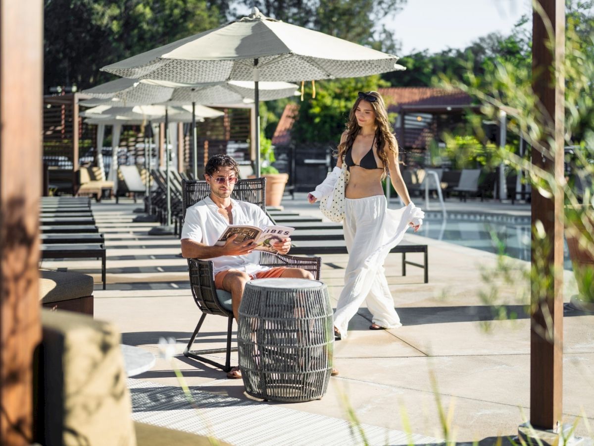 Two people relaxing by a pool area: a man seated with a drink at a small table under umbrellas, while a woman in a white skirt walks nearby.