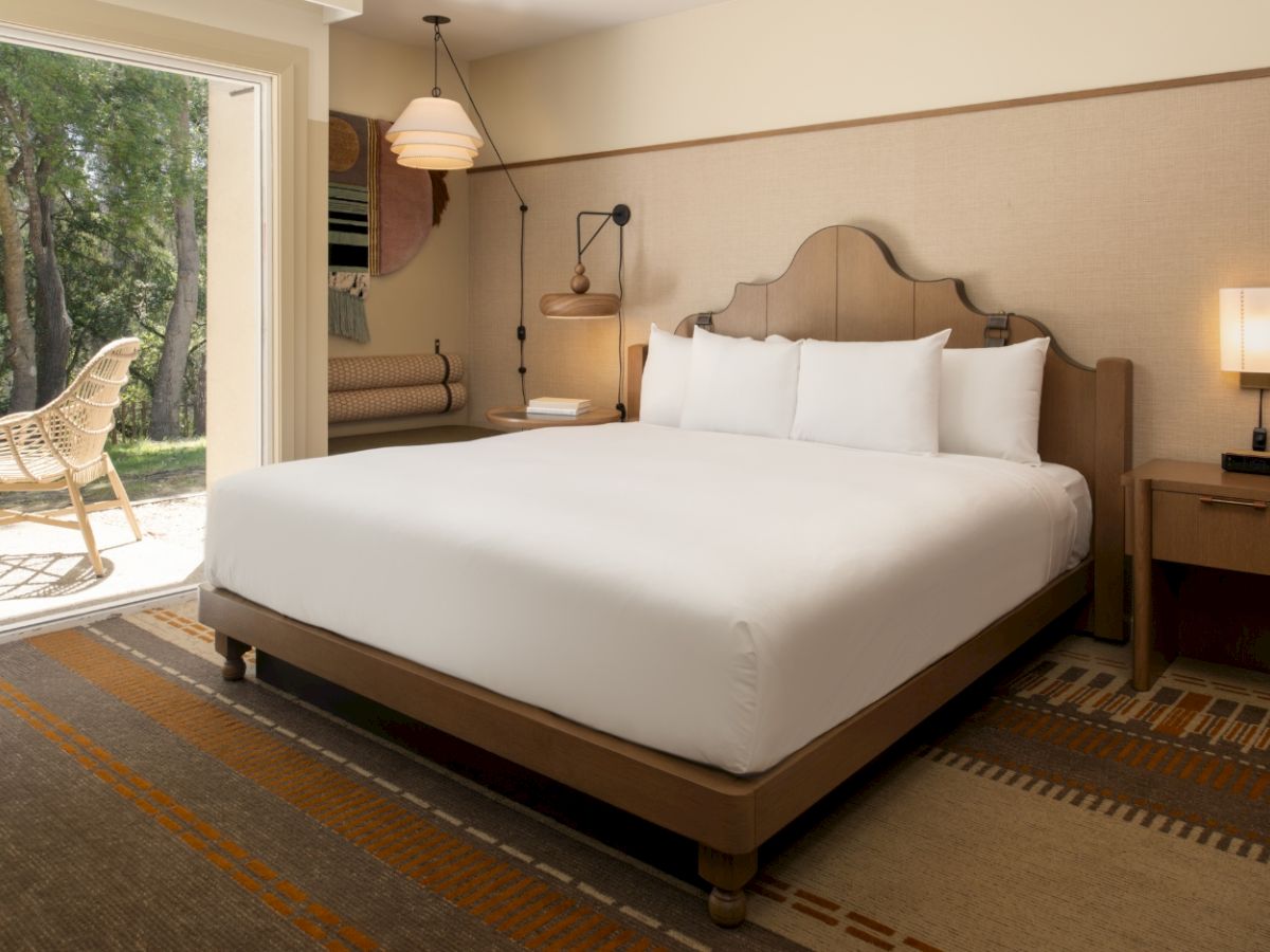 A spacious hotel room with a large bed, two nightstands, lamps, and a balcony with outdoor seating, bathed in warm light.