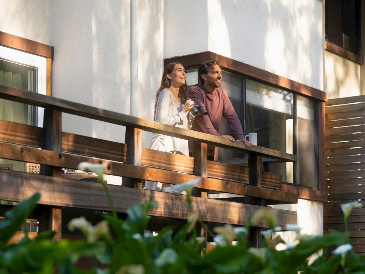 A woman and a man stand on a wooden balcony, smiling and chatting with coffee cups in warm sunlight.