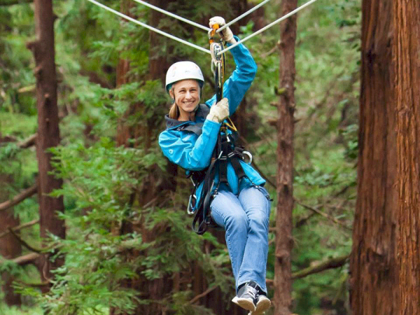 A person wearing a helmet and harness is zip-lining through a forested area with tall trees and greenery all around.