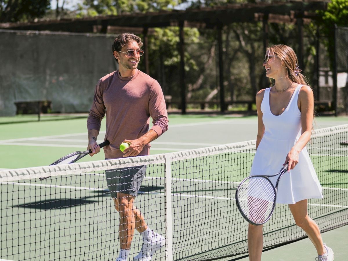 A smiling man and woman play tennis on a sunny court, holding rackets and tossing a ball back and forth.