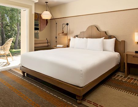 A bright hotel bedroom with a large bed, bedside lamps, patterned rug, and an open window showing trees outside.