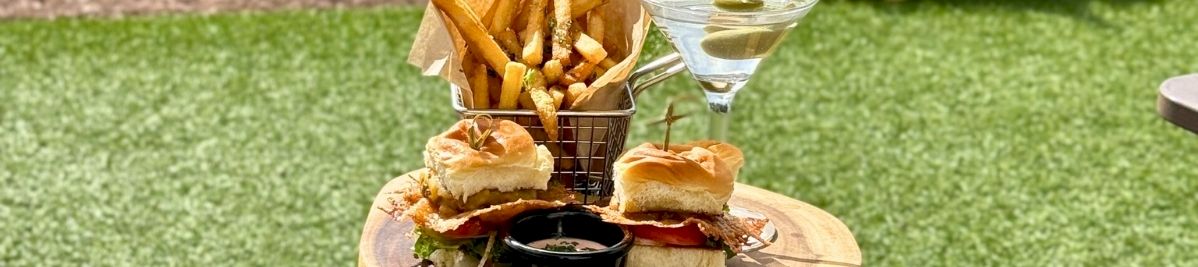 Outdoor table holds sliders, fries, dipping sauce, and a martini glass with olives in a backyard.
