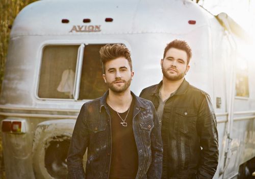 Two men stand in front of a silver trailer labeled "Avion." They both wear dark jackets and have similar hairstyles, posing outdoors in the sunlight.