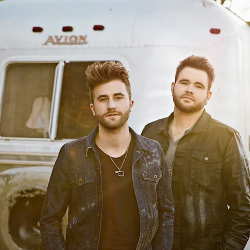 Two men stand in front of a silver trailer labeled "Avion." They both wear dark jackets and have similar hairstyles, posing outdoors in the sunlight.