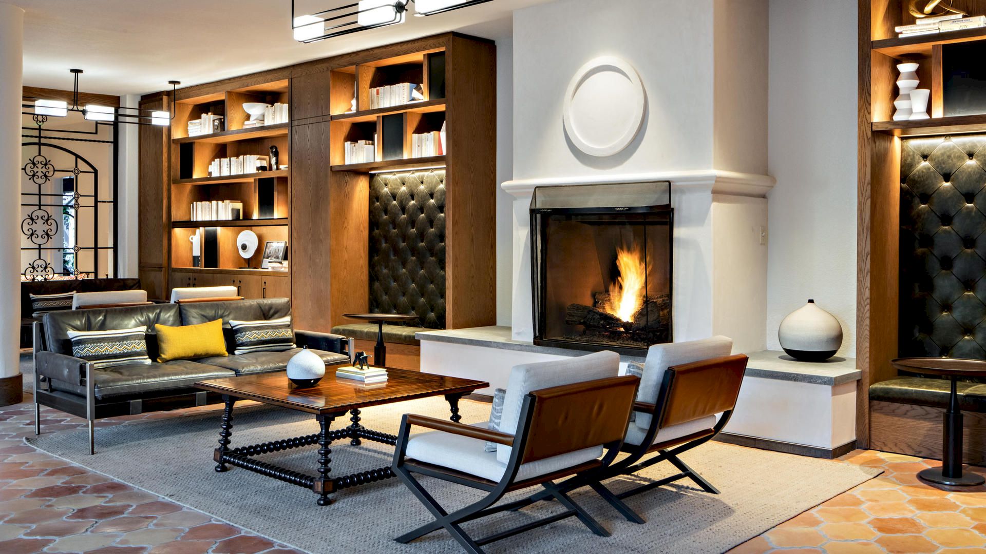 A cozy modern living room with a fireplace, leather sofa, armchairs, wooden shelves, and a coffee table, decorated with books and decor items.