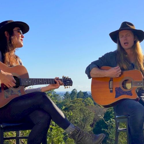 Two people sitting outdoors, playing guitars and smiling at each other, enjoying a sunny day.