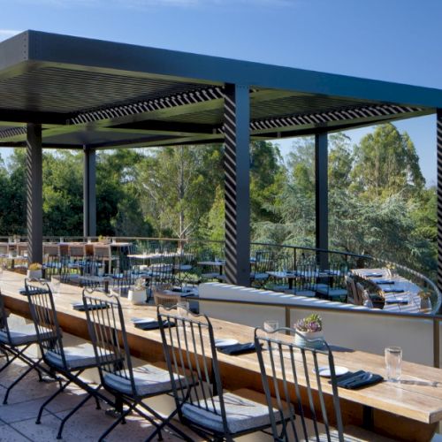 A shaded outdoor dining area with wooden tables and benches under a dark-framed pergola, overlooking a blue sky and greenery.