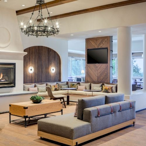 Cozy, sunlit living room with a beige sofa, a coffee table, and a striped rug; a modern fireplace, wall art, and a large window filling the space with natural light.