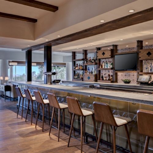 A stylish, modern cafe or bar with a long wooden counter, hanging lights, bar stools, and shelves of bottles; warm, inviting ambiance.