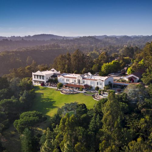 An aerial view of a large, modern house nestled among dense green trees and rolling hills, with a winding landscape and open sky.