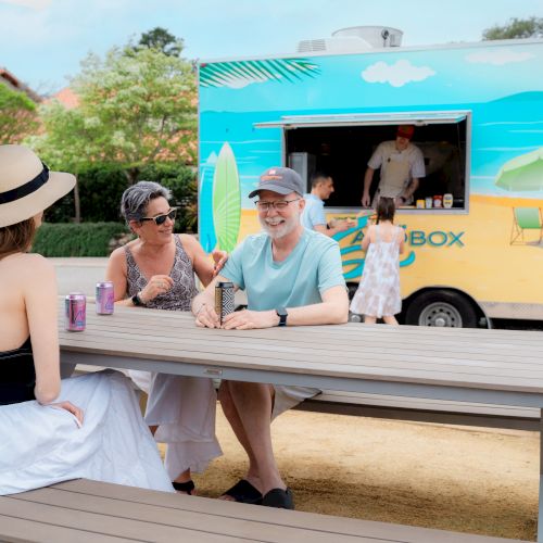 Friends enjoying drinks at a picnic bench while an ice-cream food truck serves treats in the background.