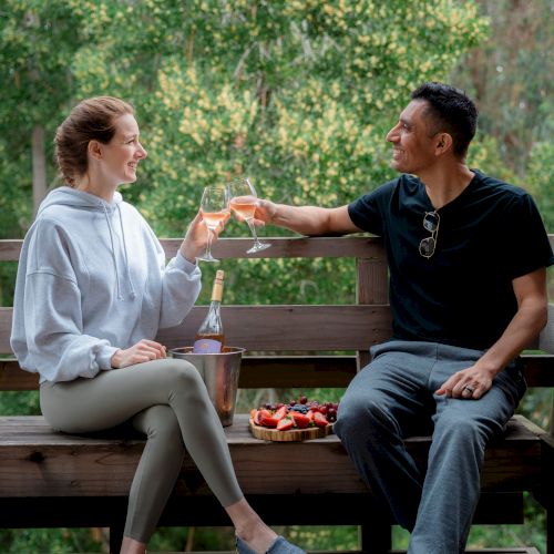 A couple clinks glasses of orange drink on a wooden bench outdoors, with fruit and a bottle between them.