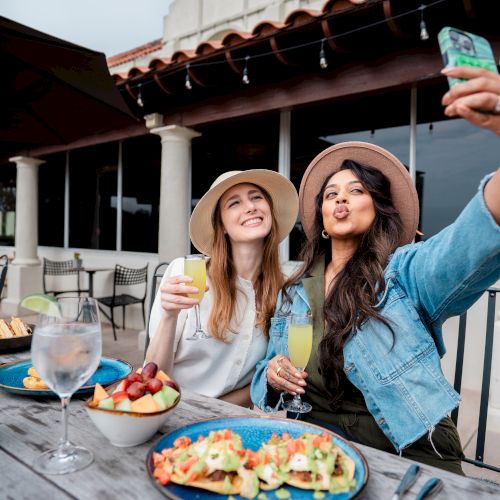 Two friends brunch on a patio, clinking glasses while one holds up a cash bill and they enjoy drinks and snacks outdoors.