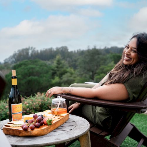 A smiling woman lounges outdoors by a picnic table with wine, fruit, and tea, set against green hills and a blue sky.