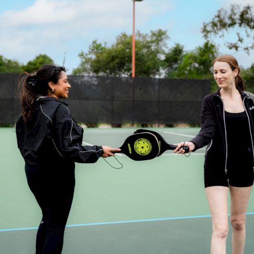Two women on an outdoor green court trade rackets and smiles during a friendly match.