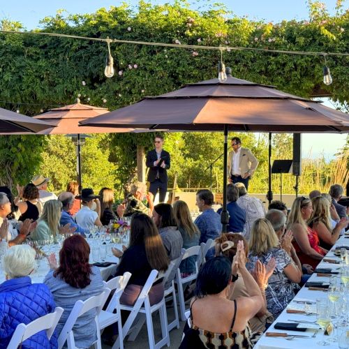 A lively outdoor gathering with people seated at long tables under tents, enjoying food and conversation at a sunny event.