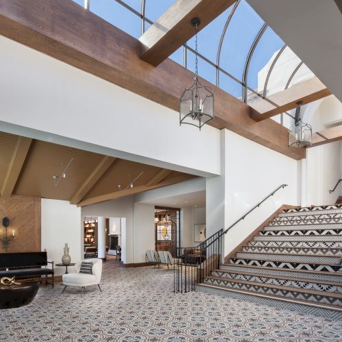 A covered outdoor walkway or corridor with a wooden railing, columns, and sunlight streaming in, leading to a stairwell. Top it at 140 characters, always ending the sentence.
