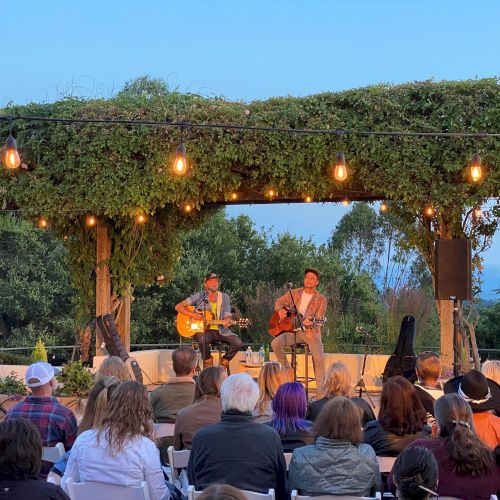 Outdoor wedding or ceremony on a sunny day with guests seated, greenery, and a wooden arch; a small crowd enjoying the event.