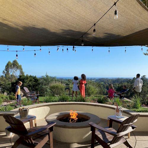 A sunny outdoor dining setup with a long wooden table, chairs, and people relaxing by a lake or river in a park-like area, mountains in the distance.