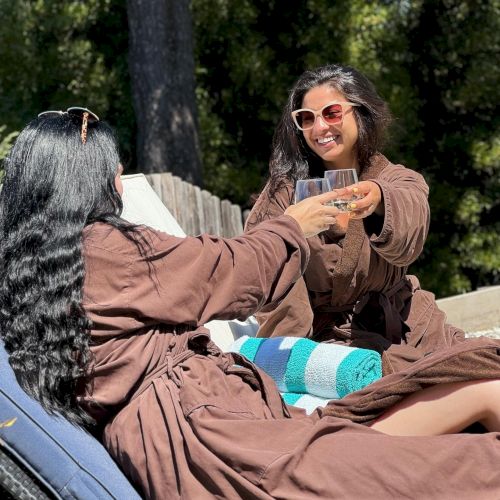Two hikers rest on a camping tarp in a forest, one person sitting in a brown robe-like blanket, another partially visible, with gear around them.