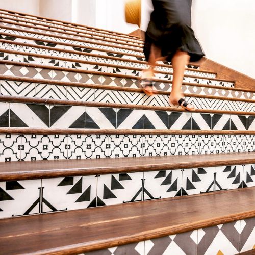 A person sits with legs crossed on patterned woven rugs or textiles, featuring geometric black and brown motifs.