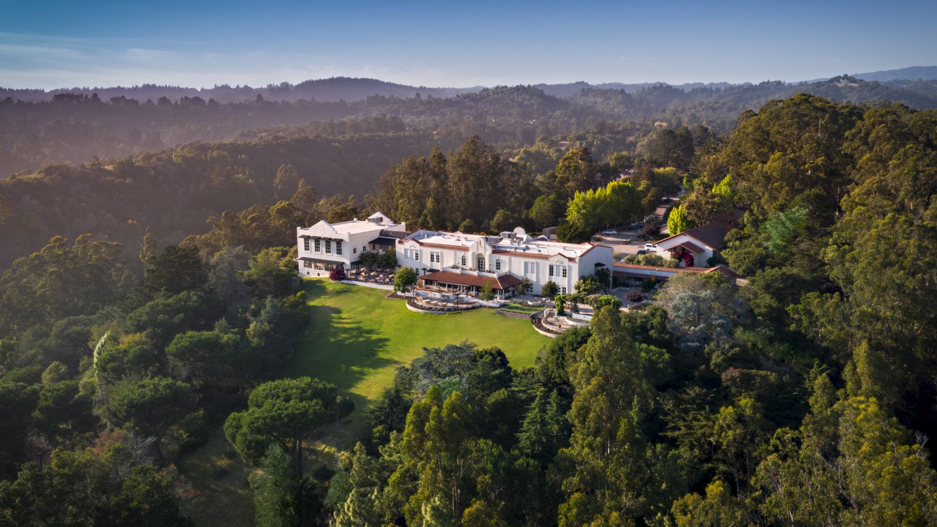Aerial view of a white villa with red-tile roofs surrounded by lush greenery and rolling hills at golden-hour light.