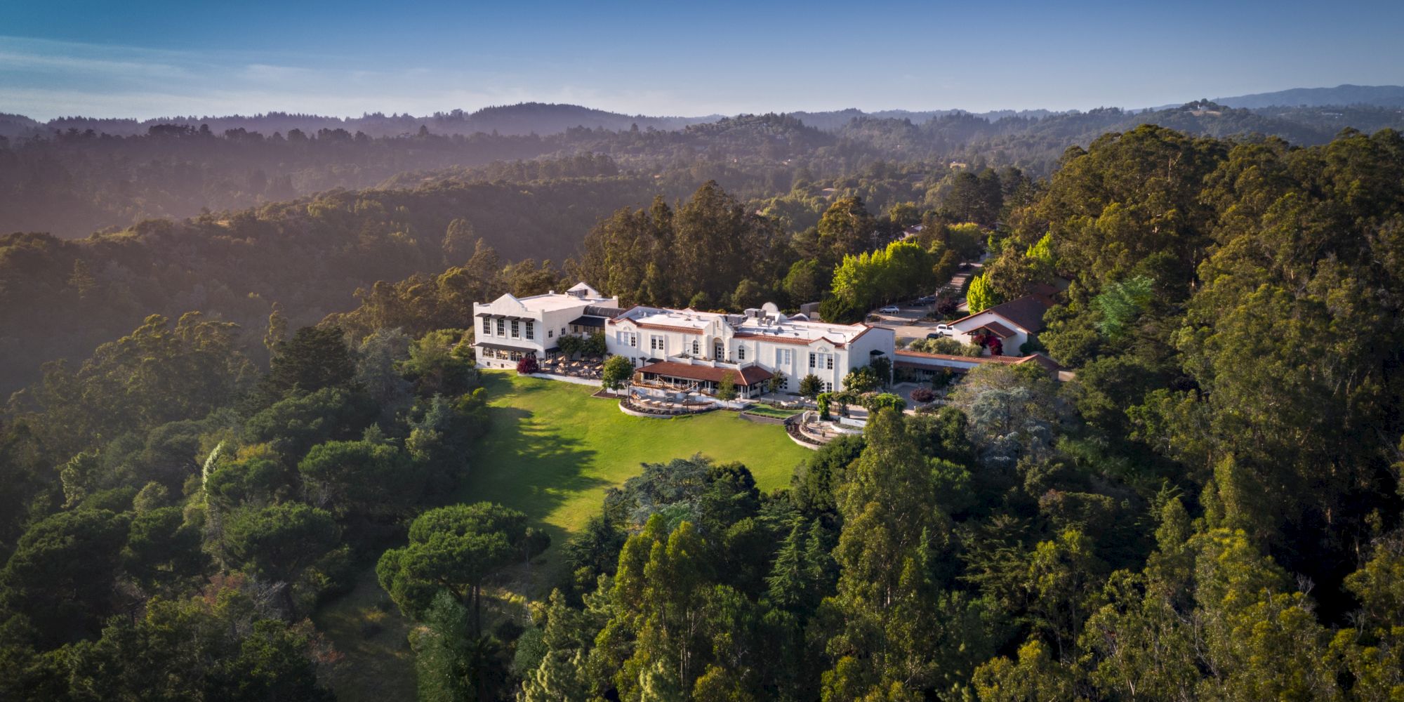 Aerial view of a white villa with red-tile roofs surrounded by lush greenery and rolling hills at golden-hour light.