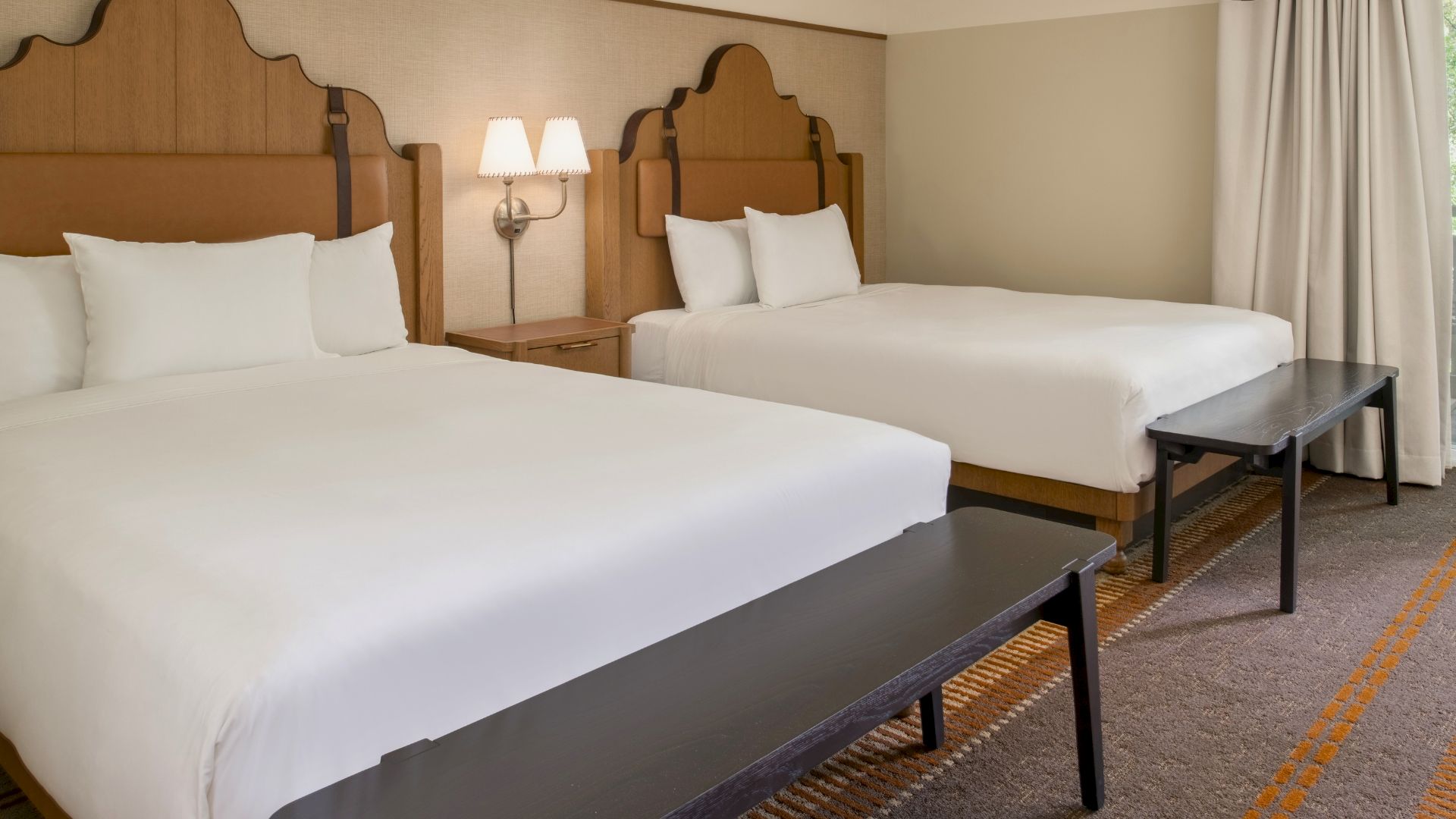 Two queen beds with white linens, wooden headboards, a shared nightstand with a lamp, and a bench at the foot of each bed in a hotel room.