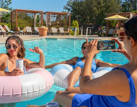 Friends lounge in a sunny outdoor pool on inflatable rings, posing and waving while someone snaps a phone photo.