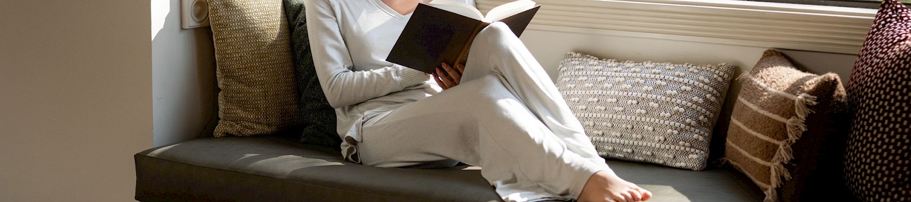 A woman lounges on a cushioned window bench reading a book in warm sunlight, with pillows and a camera on a table.