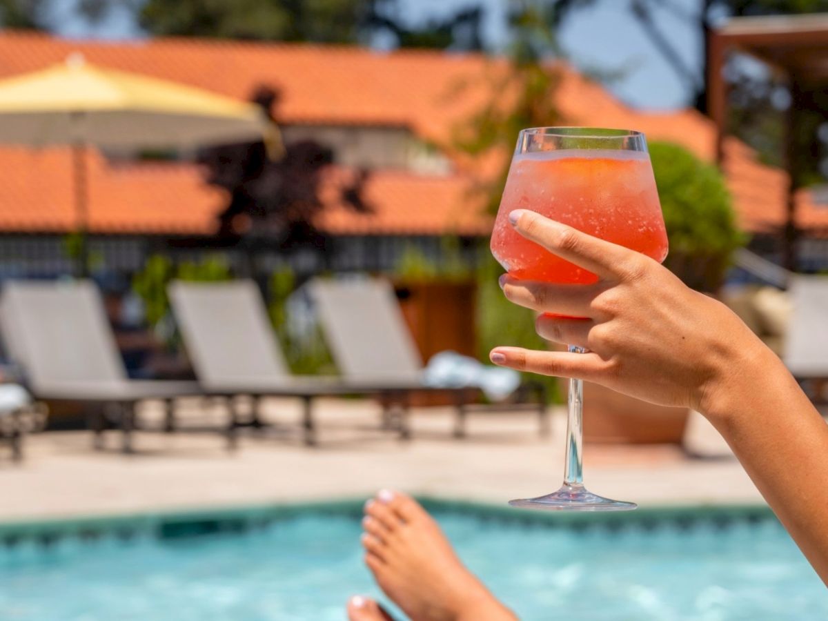 A hand lifts a pink drink near a sunny pool with lounge chairs behind it.