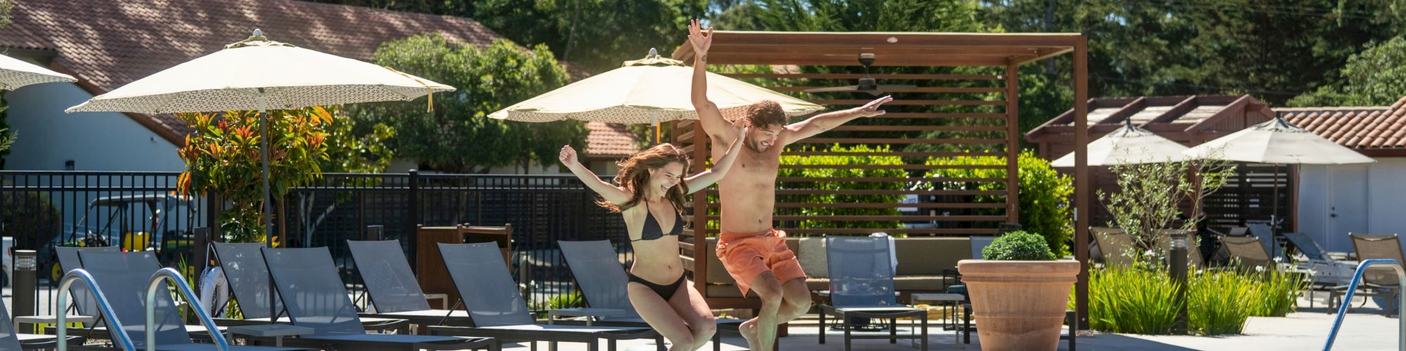 A couple jumps into a sunny pool at a resort, arms out and splashing near loungers and umbrellas.