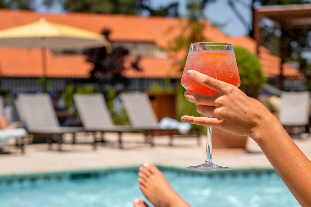 A hand holds a pink cocktail in a glass beside a sunny outdoor pool.