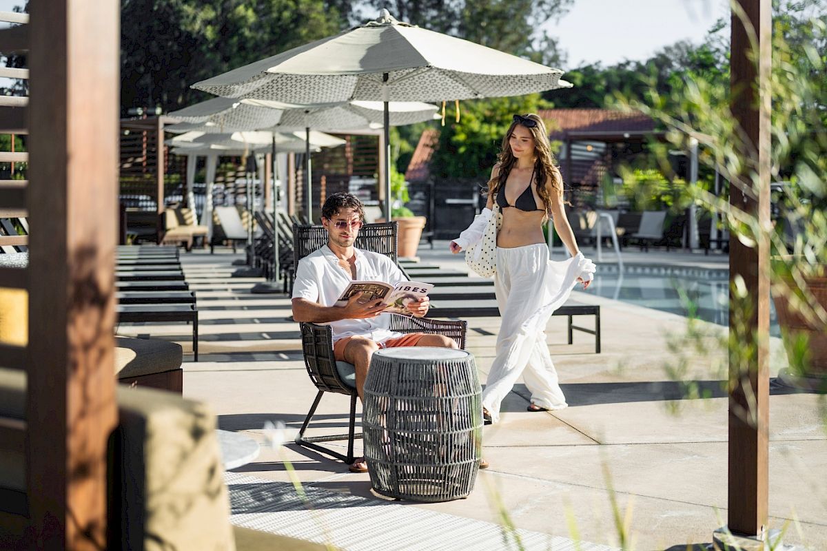 An outdoor resort scene: a man lounges reading on a chair while a woman in a bikini and white robe dances near umbrellas.