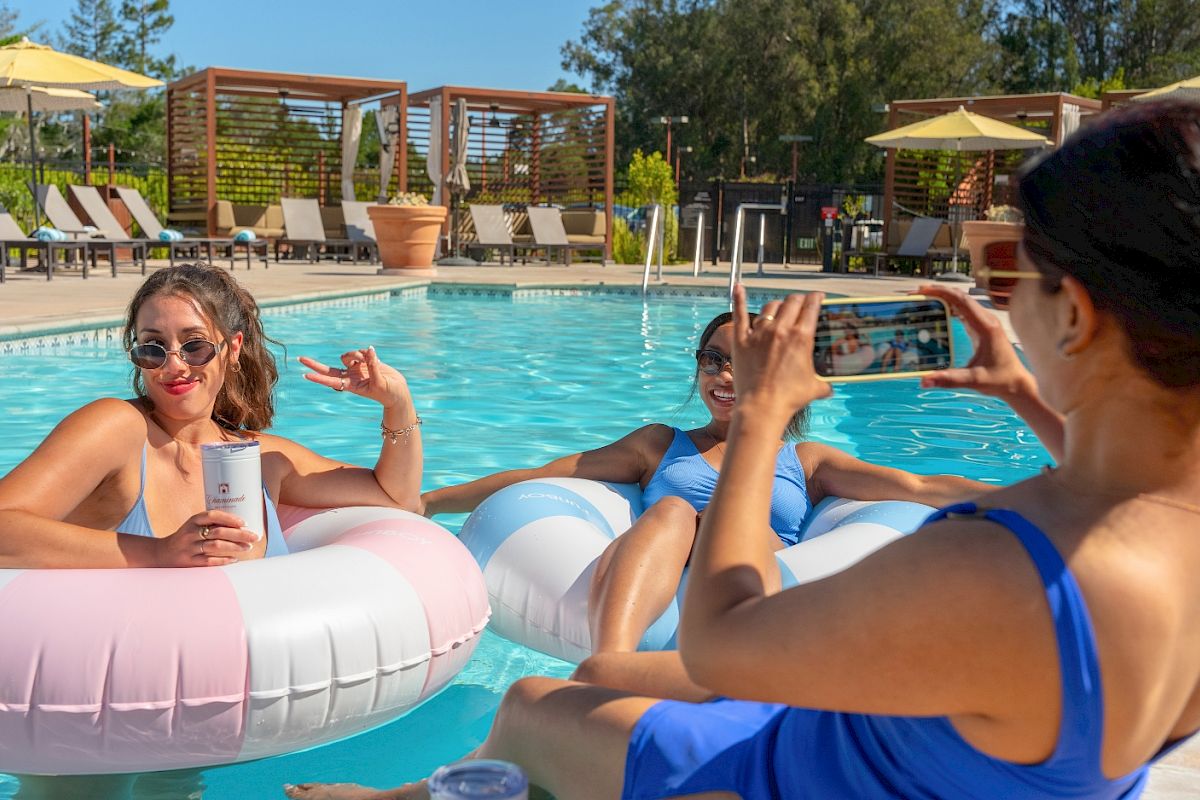 Friends lounge in the pool on inflatable floats at a sunny resort, smiling and waving as someone takes a photo.