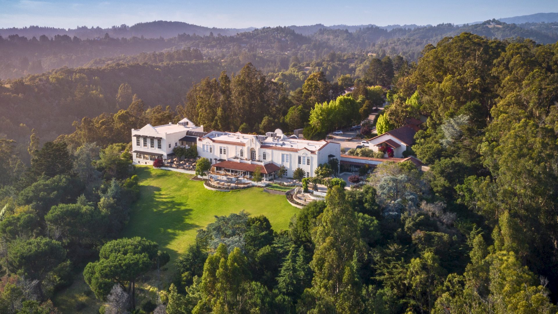 Aerial view of a white Mediterranean-style mansion with terraced gardens, surrounded by lush trees and rolling hills.